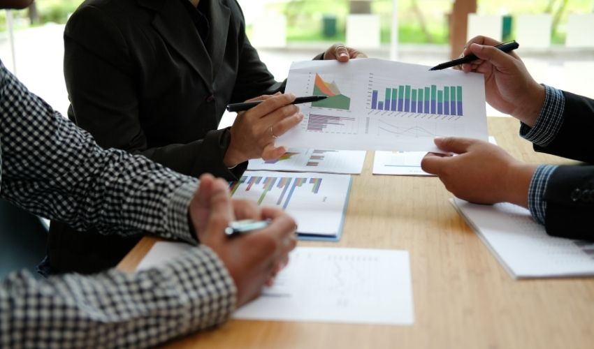 Three people sitting at a table review printed charts and graphs, discussing data related to kanada-experten-migration while pointing at the documents with pens. The setting appears to be a business meeting or discussion.